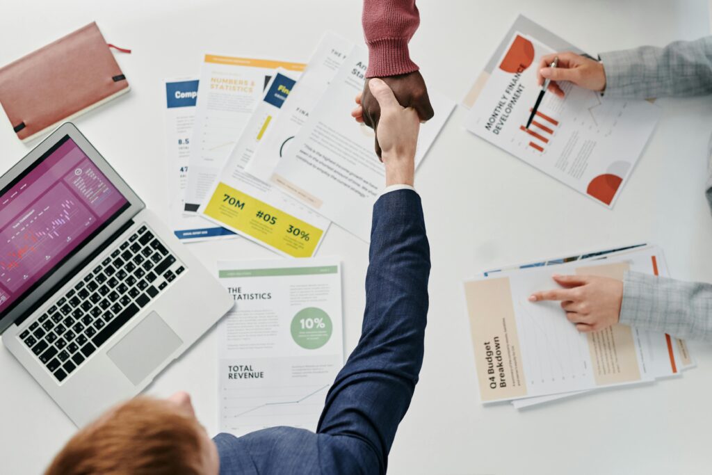 Business professionals engaging in a handshake over a desk filled with financial documents and a laptop.